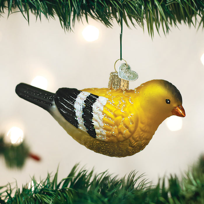 A yellow and black bird-shaped Christmas ornament hangs from a green tree branch, with soft, out-of-focus white lights in the background.