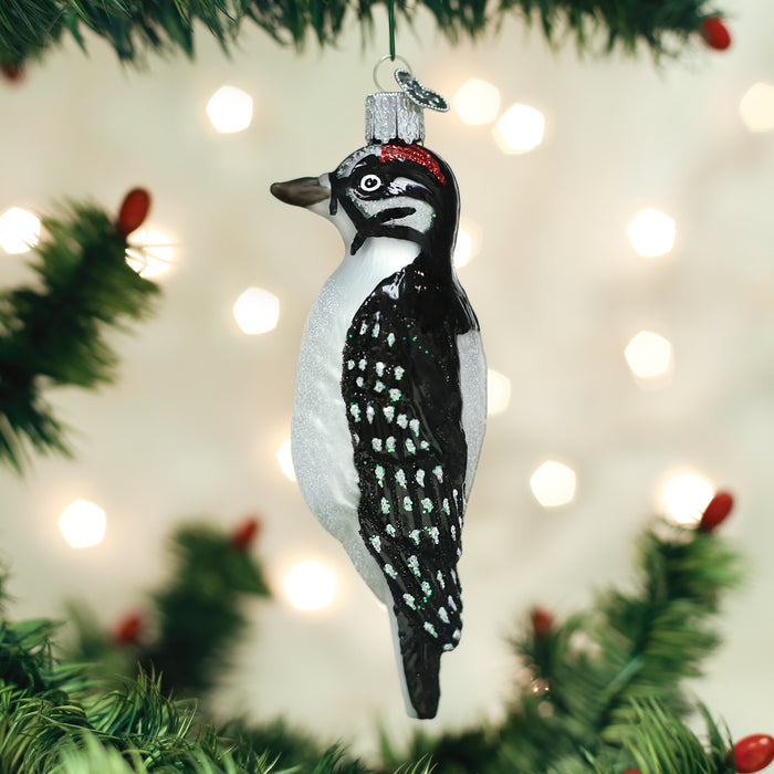 A glass ornament shaped like a woodpecker with black, white, and red markings hangs from a green Christmas tree branch, with blurred white lights in the background.