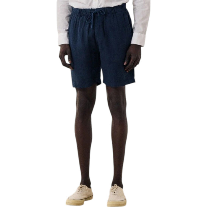 A person wearing a white long-sleeve shirt, HARTFORD - LINEN JOGGY SHORT IN NAVY by HARTFORD, and beige sneakers stands on a wooden floor against a plain light background.