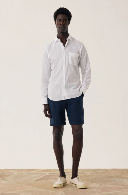 A person wearing a white long-sleeve shirt, HARTFORD - LINEN JOGGY SHORT IN NAVY by HARTFORD, and beige sneakers stands on a wooden floor against a plain light background.