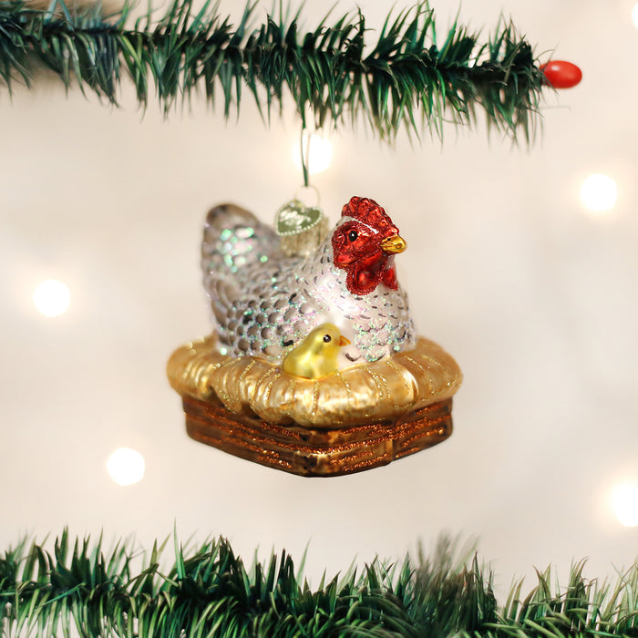 A glass Christmas tree ornament shaped like a white hen with a red comb and a yellow chick, both sitting on a nest, hangs on a green tree branch with white lights in the background.