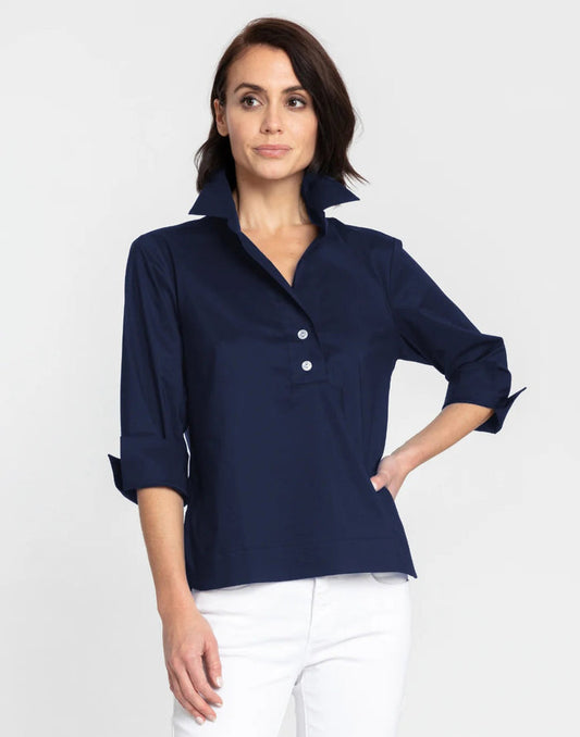 A woman with short dark hair models the HINSON WU - AILEEN 3/4 SLEEVE POLISHED COTTON SHIRT in navy, featuring a collared neckline, stretch fabric, and three buttons. She pairs it with white pants and poses against a plain light gray background.