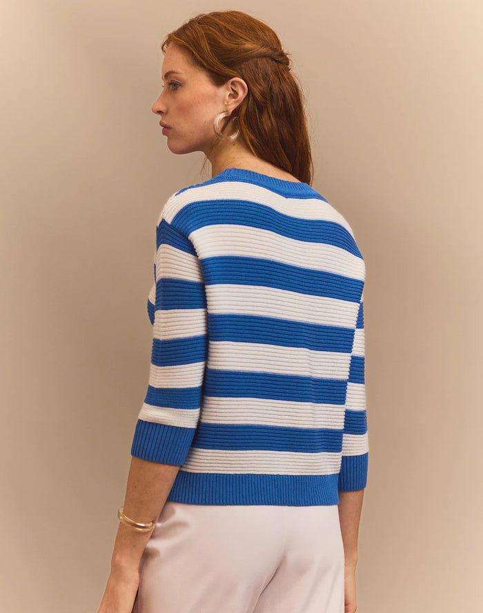 A woman with long red hair wears the HINSON WU CIARA 3/4 Sleeve Stripe Sweater and white pants, holding a large woven straw handbag, standing against a beige background.