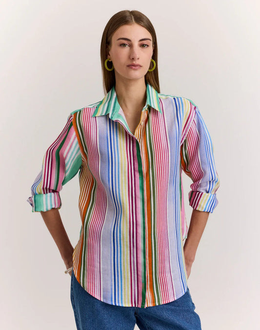 A woman in the HINSON WU Halsey Long Sleeve Linen Shirt in Multi Stripe, featuring vibrant vertical stripes, rolled-up sleeves, and lightweight linen fabric, pairs it with blue jeans. She has straight brown hair, hoop earrings, and a bracelet.