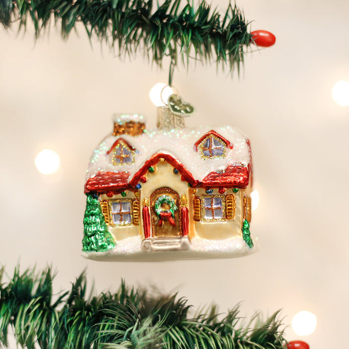 A glass Christmas ornament shaped like a snowy house with red roof, wreath on the door, and green trees hangs on a decorated Christmas tree branch with blurred lights in the background.