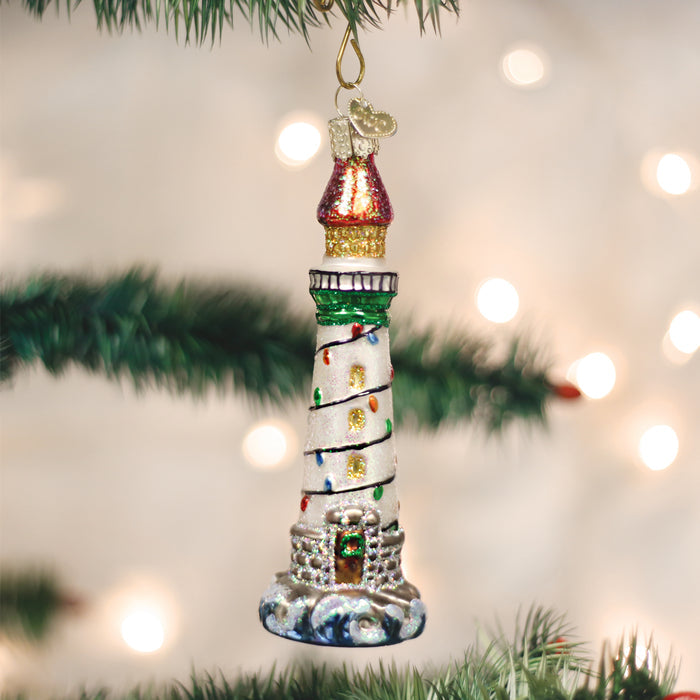 A glass lighthouse ornament decorated with colorful lights hangs from a Christmas tree branch, with soft white holiday lights blurred in the background.