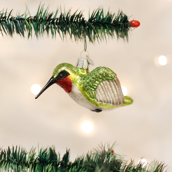 A glass hummingbird ornament with green wings and a red throat hangs from a Christmas tree branch, with blurred white lights in the background.