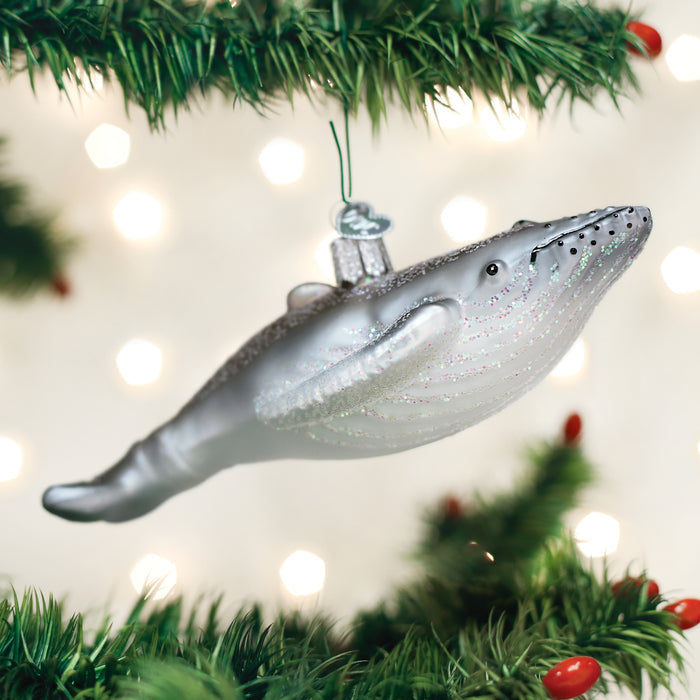 A silver whale ornament hangs from a Christmas tree branch, surrounded by green pine needles and blurred white holiday lights in the background.
