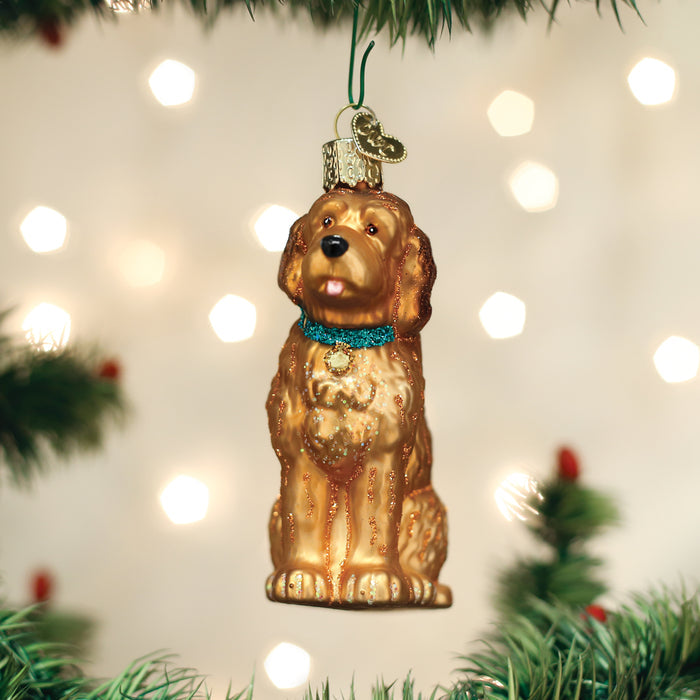A golden dog-shaped Christmas ornament with a blue collar hangs from a tree branch, surrounded by green pine needles and festive lights in the background.