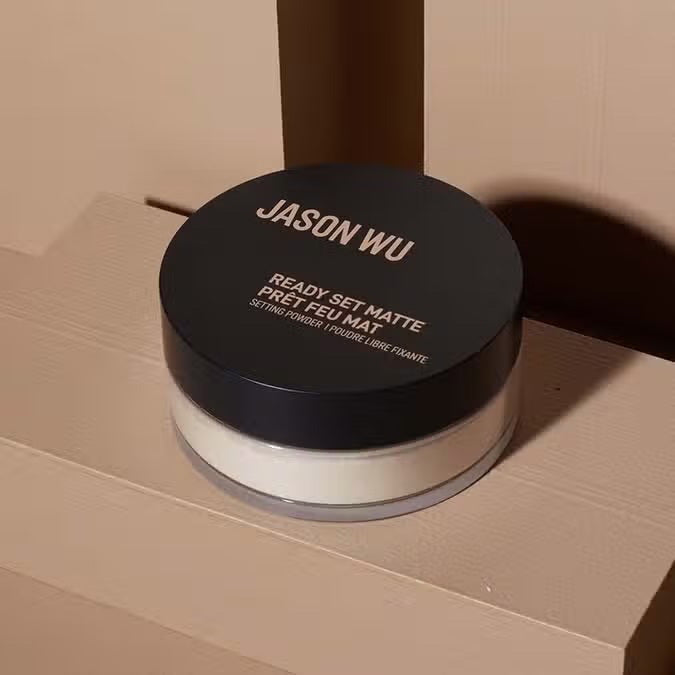 A round container of JASON WU BEAUTY READY SET MATTE 1 - TRANSLUCENT BANANA setting powder with a black lid and beige powder rests on a light brown textured surface.