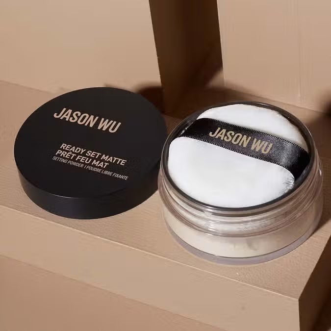 A round container of JASON WU BEAUTY READY SET MATTE 1 - TRANSLUCENT BANANA setting powder with a black lid and beige powder rests on a light brown textured surface.