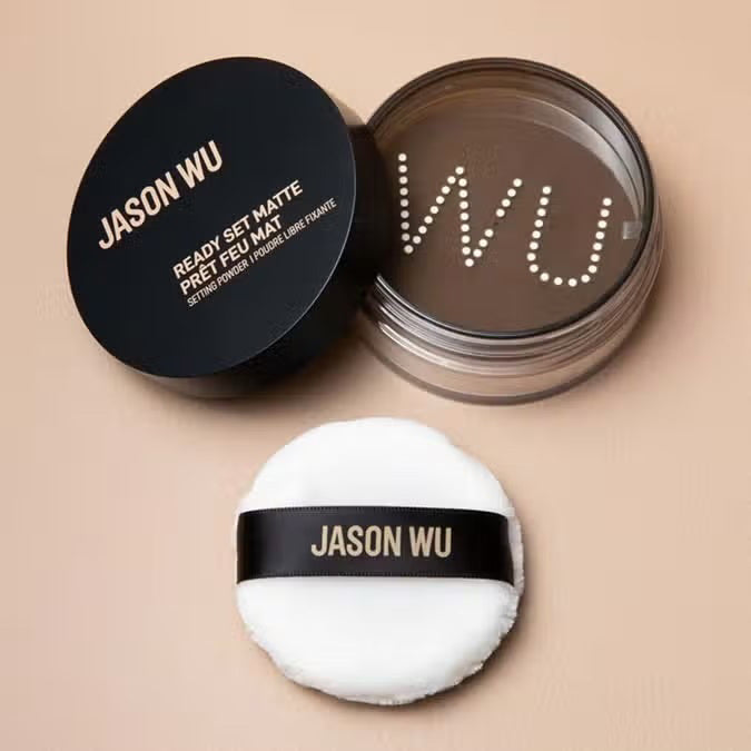 A round container of JASON WU BEAUTY READY SET MATTE 1 - TRANSLUCENT BANANA setting powder with a black lid and beige powder rests on a light brown textured surface.