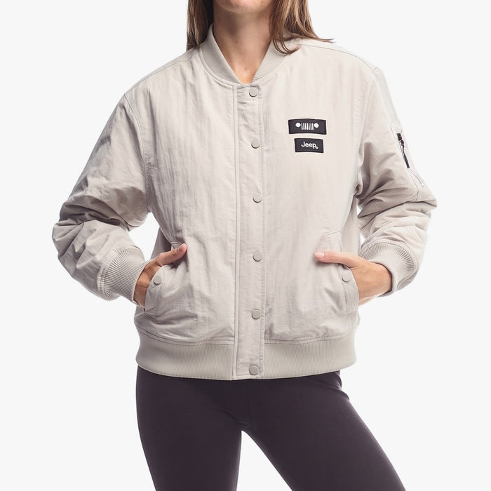 Woman wearing a khaki Jeep bomber jacket with ribbed cuffs and hem, featuring a black Jeep logo on the chest. Hands are in