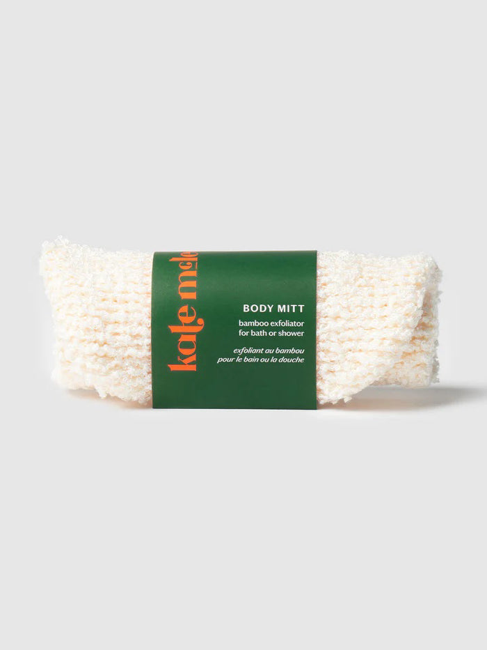 A hand showcases a textured beige KATE MCLEOD - THE BODY MITT against a light turquoise background, while another hand adjusts the cuff with its black tag and red stitching. Beneath, the white surface featuring gray veining creates an ideal exfoliating spa treatment ambiance.