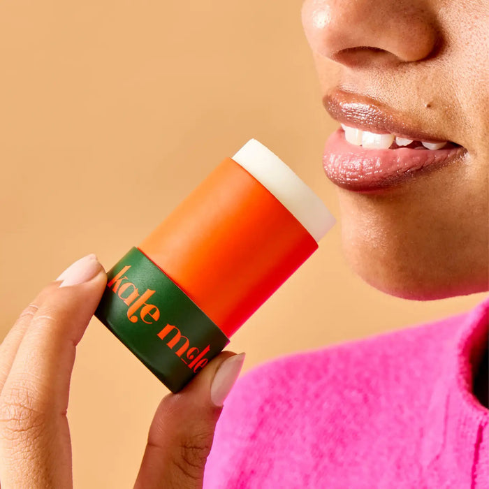 A cylindrical skincare stick features an orange design with "KATE MCLEOD" inscribed. Next to it, a green cap displays "The Everywhere Stick" in white. A red circle above says "New In." Made with pure plant butters, this wax-free formula is a hydration hero, all set against a light green background.