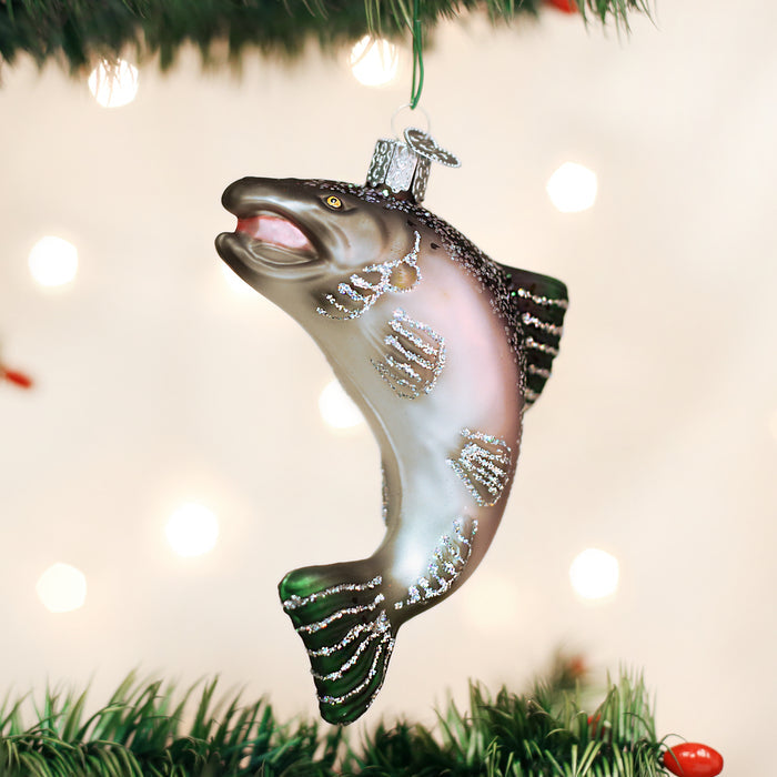 A glass ornament shaped and painted like a leaping fish hangs on a Christmas tree, with blurred white lights in the background.