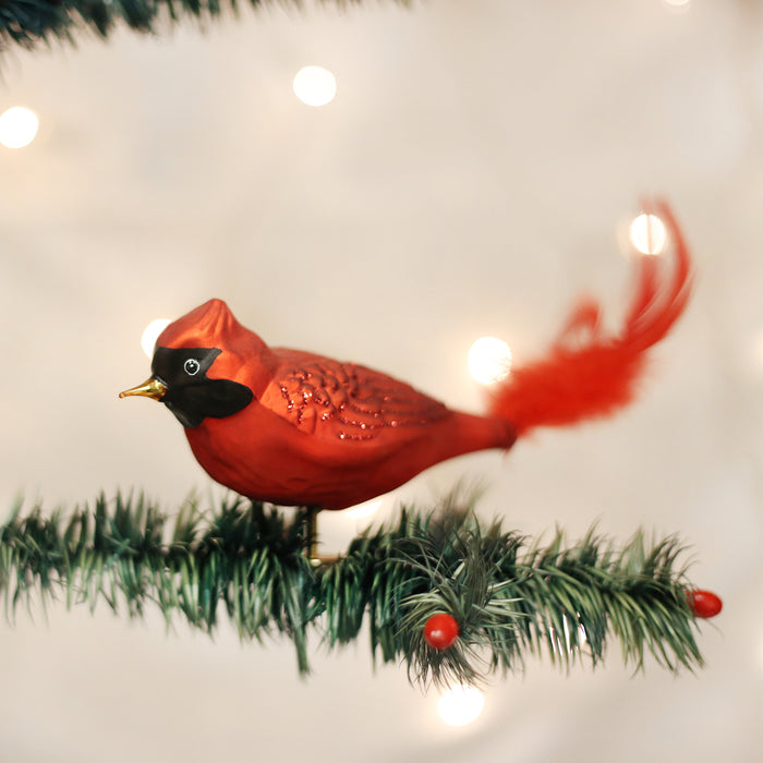 A red cardinal ornament with a feathered tail sits on a green artificial pine branch, with white holiday lights softly glowing in the blurred background.