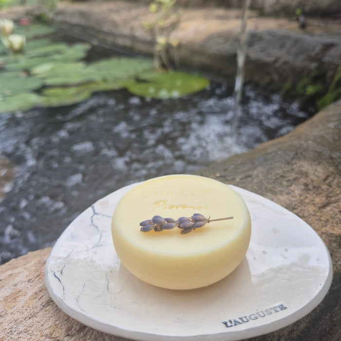 The L’AUGUSTE PROVENCE - LAVENDER SOAP AND ARTISANAL SOAP DISH by L’AUGUSTE PROVENCE sits on stone near a pond, with the round engraved soap topped by lavender and displayed on a white dish.