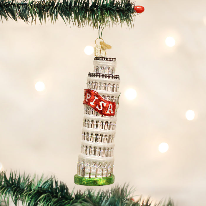 A Christmas tree ornament shaped like the Leaning Tower of Pisa hangs from a green tree branch. The ornament is decorated with glitter and the word PISA in red letters. Soft white lights glow in the background.