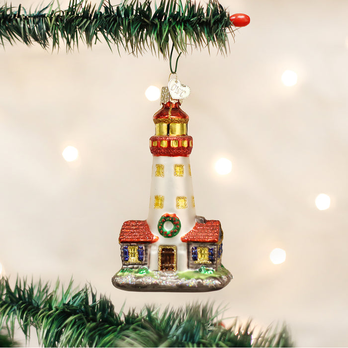 A glass Christmas ornament shaped like a lighthouse with a wreath on the door hangs from a green tree branch, with soft white holiday lights in the blurred background.