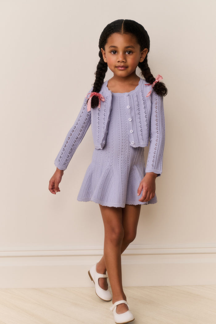 Young girl wearing a light blue cardigan and dress outfit against a plain background