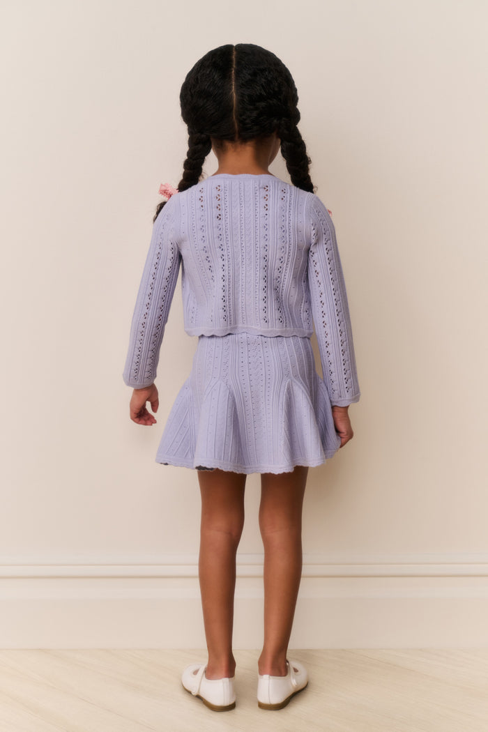 Young girl wearing a light blue cardigan and dress outfit against a plain background
