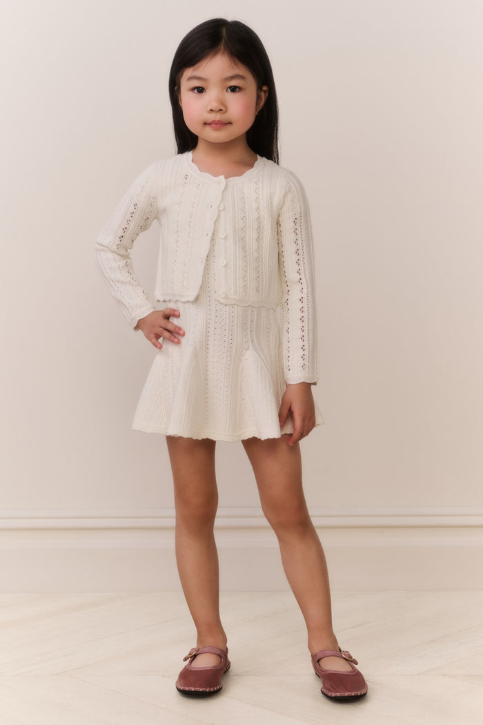 Young girl wearing a cream-colored knit cardigan and skirt outfit against a plain background