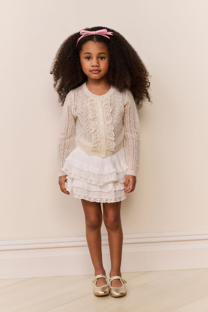 Young girl wearing a white ruffle cardigan and skirt with a pink bow in her hair against a plain background
