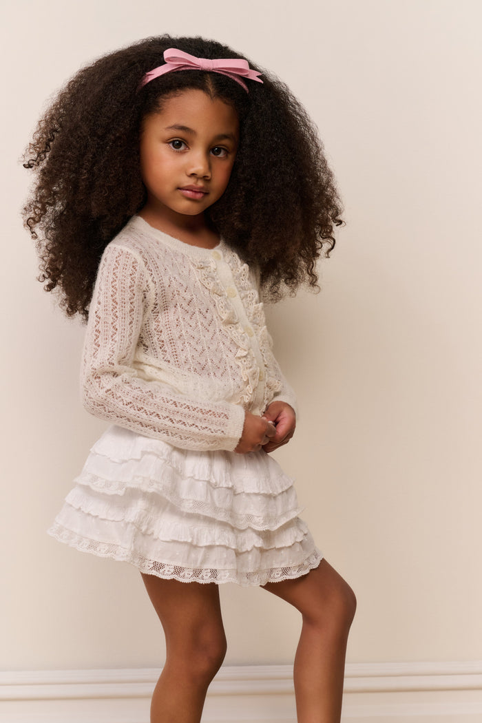 Young girl wearing a white ruffle cardigan and skirt with a pink bow in her hair against a plain background