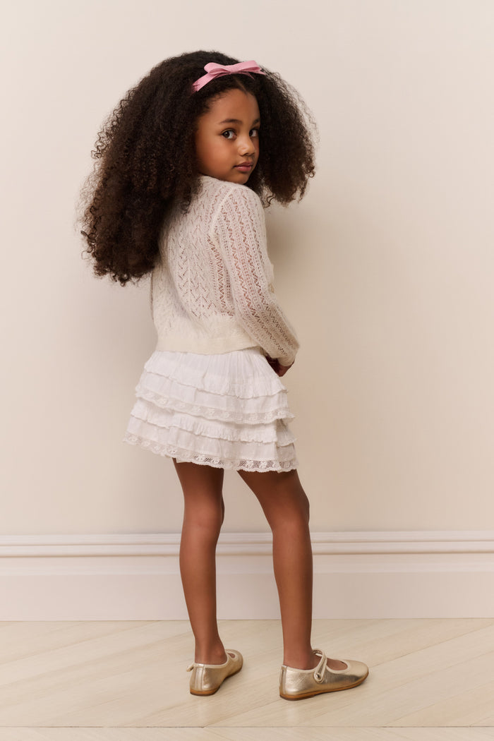 Young girl in a white cardigan and dress with a pink bow standing against a plain wall.