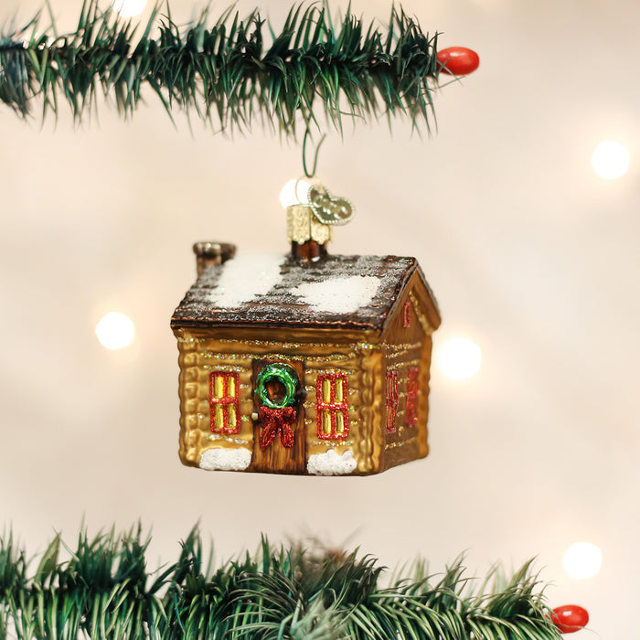 A glass Christmas ornament shaped like a small snow-covered house hangs from a green tree branch, with festive decorations and blurred lights in the background.