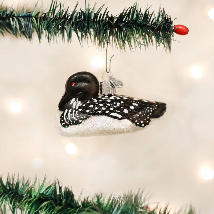 A Christmas tree branch holds a glass ornament shaped like a black and white loon. The background is softly blurred with warm white holiday lights.