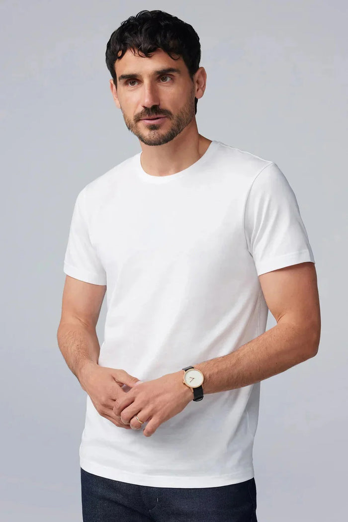 A man with short dark hair and a beard wears the Mack Weldon Silver Crew Neck T-Shirt in Desert Taupe, paired with dark jeans and a wristwatch. He stands against a light gray background, looking slightly to the side with a neutral expression.