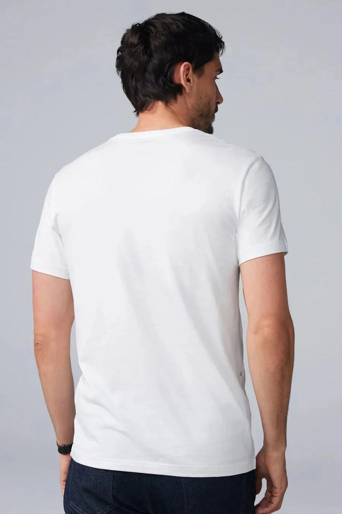 A man with short dark hair and a beard wears the Mack Weldon Silver Crew Neck T-Shirt in Desert Taupe, paired with dark jeans and a wristwatch. He stands against a light gray background, looking slightly to the side with a neutral expression.