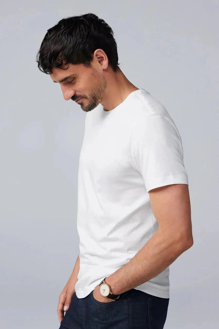 A man with short dark hair and a beard wears the Mack Weldon Silver Crew Neck T-Shirt in Desert Taupe, paired with dark jeans and a wristwatch. He stands against a light gray background, looking slightly to the side with a neutral expression.