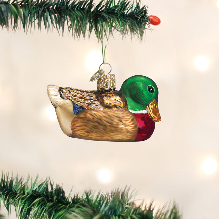 A glass Christmas tree ornament shaped and painted like a mallard duck hangs from a green pine branch, with soft, blurred white lights in the background.