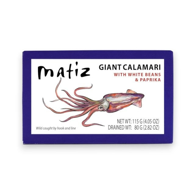 A bowl of rice salad with vegetables and wild-caught calamari sits beside a box labeled “Matiz España - Giant Calamari with White Beans and Paprika,” a small bowl of this gourmet seafood from Matiz España, a wooden fork and spoon, and a dish of green sauce infused with Spanish paprika on a white table.