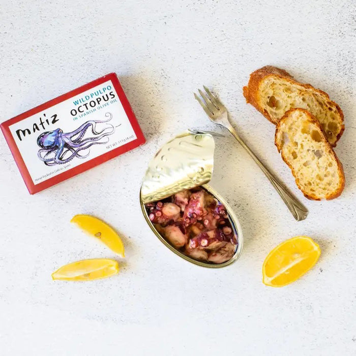A rectangular tin can with a red border and white label. The label features an illustration of a purple octopus and text that reads "MATIZ - PULPO (OCTOPUS) IN OLIVE OIL" by MATIZ ESPANA and "Hand-Packed in Galicia, Spain." The net weight is 111g (4 oz).