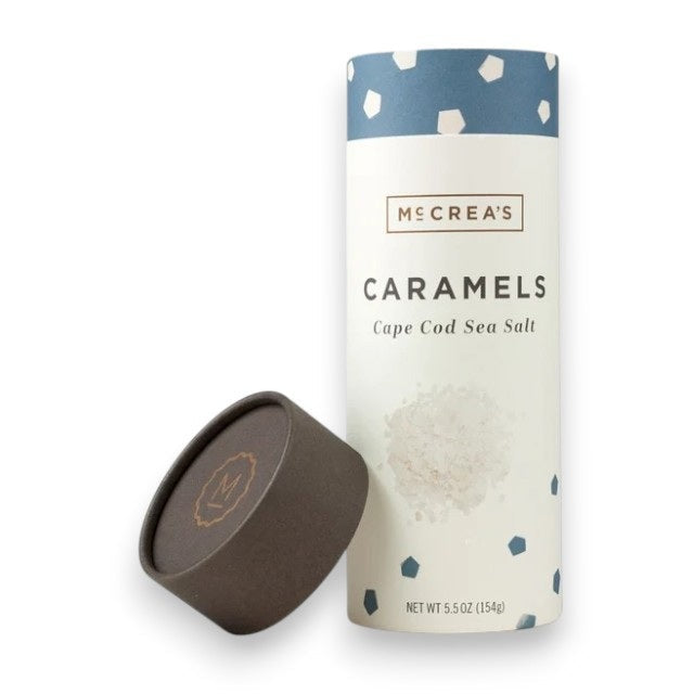 A cylindrical container of McCREA’S CANDIES - CAPE COD SEA SALT CARAMELS, featuring a blue and white geometric design inspired by classic New England charm, with the lid removed and placed beside it. Net weight: 5.5 oz (154g).