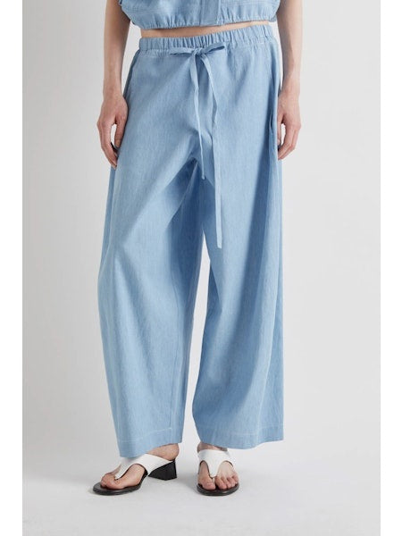 Model wears MEIMEIJ - DRAWSTRING COTTON PANTS in light blue, featuring a loose fit, wide legs, cropped ankle, and drawstring waist. Styled with white sandals. Image shows shoulders to feet against a plain background.