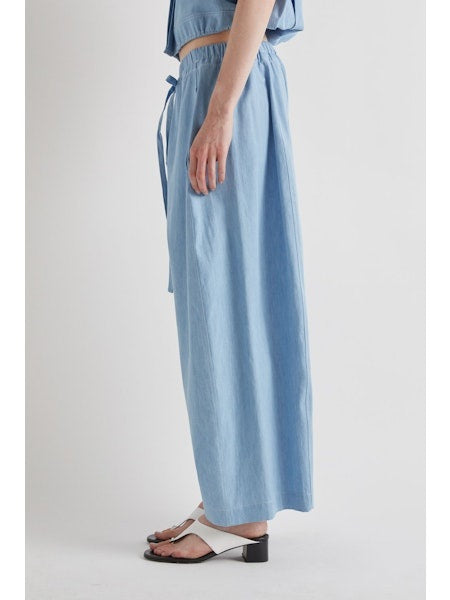 Model wears MEIMEIJ - DRAWSTRING COTTON PANTS in light blue, featuring a loose fit, wide legs, cropped ankle, and drawstring waist. Styled with white sandals. Image shows shoulders to feet against a plain background.