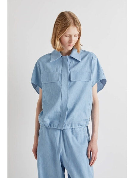A person with chin-length light brown hair stands with their back to the camera, wearing the MEIMEIJ DRAWSTRING COTTON TOP in chambray blue. The loose fit and plain white background highlight MEIMEIJ's modern Italian aesthetic.