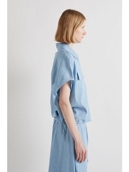 A person with chin-length light brown hair stands with their back to the camera, wearing the MEIMEIJ DRAWSTRING COTTON TOP in chambray blue. The loose fit and plain white background highlight MEIMEIJ's modern Italian aesthetic.