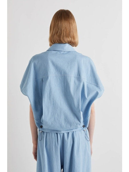 A person with chin-length light brown hair stands with their back to the camera, wearing the MEIMEIJ DRAWSTRING COTTON TOP in chambray blue. The loose fit and plain white background highlight MEIMEIJ's modern Italian aesthetic.