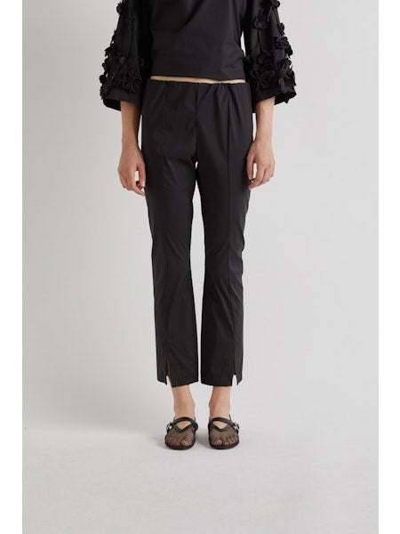 A person stands against a plain background wearing MEIMEIJ FROTN HEM SPLIT COTTON PANT—black, cropped, ankle-length pants with small slits. They also wear a black textured-sleeve top and black shoes. Their face is not visible in the image.