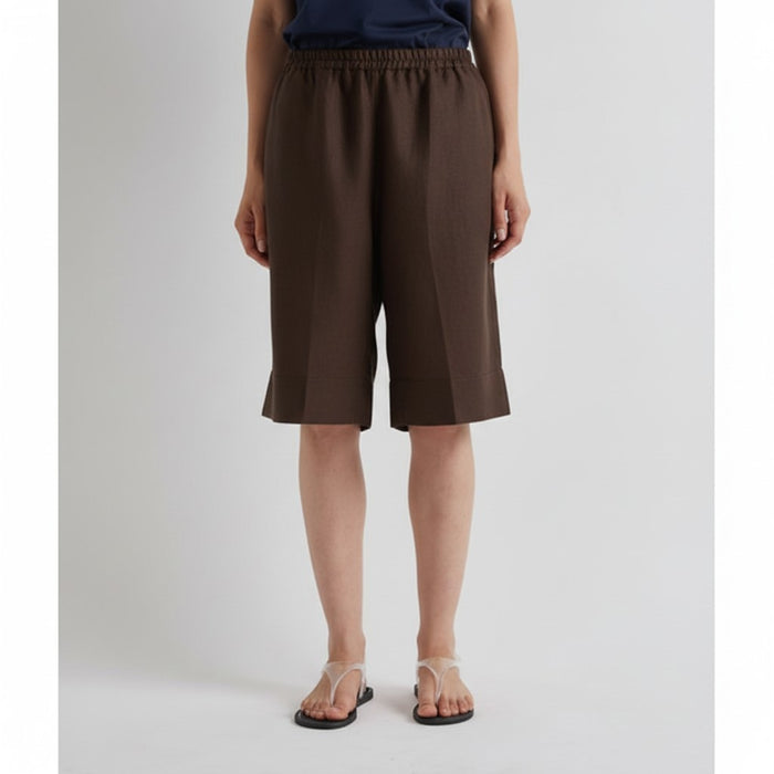 A person wears MEIMEIJ's Linen Shorts in Mocha and clear strap sandals with a navy top, standing against a light background, embodying relaxed summer tailoring.