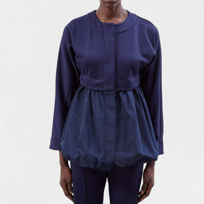 A person stands against a plain background wearing the MEIMEIJ - PONTE KNIT AND TAFFETA JACKET in navy blue. The fitted upper section transitions into loose, gathered fabric starting at the midsection, creating a unique silhouette reminiscent of taffeta bottoms. Matching navy blue pants complete the look from MEIMEIJ.