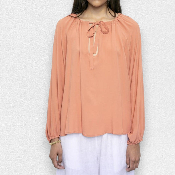A woman in the MEIMEIJ Tie Neck Blouse—loose, long-sleeved, peach-colored washable silk with a neckline tie—paired with white pants, stands against a light textured background. Her face is partially out of frame.