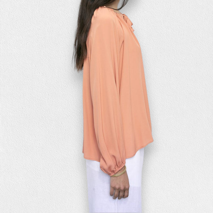 A woman in the MEIMEIJ Tie Neck Blouse—loose, long-sleeved, peach-colored washable silk with a neckline tie—paired with white pants, stands against a light textured background. Her face is partially out of frame.