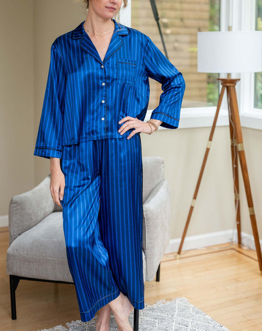 The MERSEA - Carousel Stripe Satin Sailor Pajama Set in Blue by MERSEA, featuring elegant blue and black stripes with a button-up top and matching pants, embodies luxury sleepwear and is neatly folded on a light blue background.